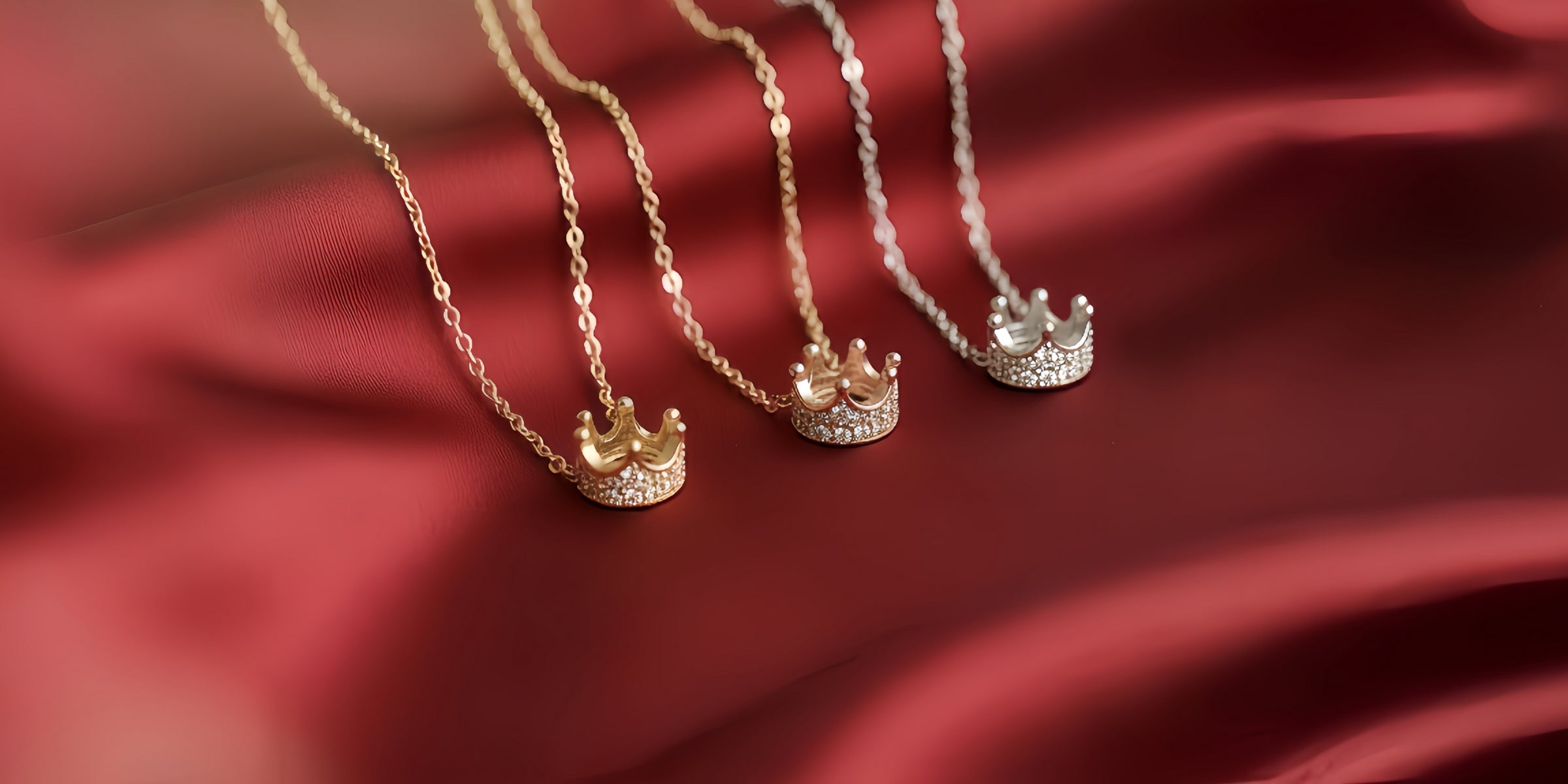 Three crown pendant necklaces on red satin fabric, shown in gold, rose gold and silver tones.