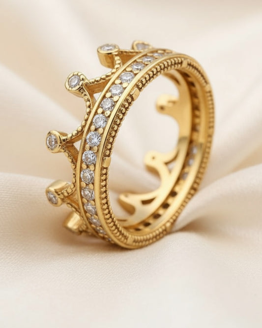 Close-up of a gold crown-shaped ring with clear stones, resting on soft ivory satin fabric.