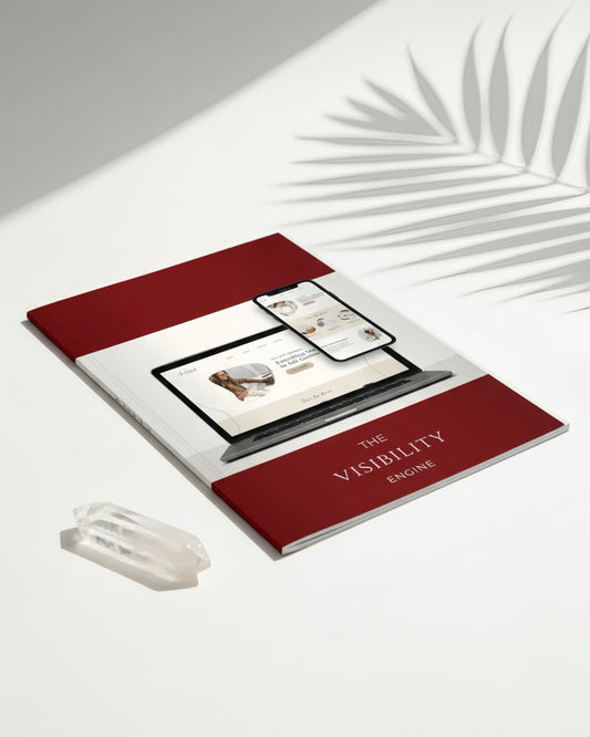 Pamela Blaney’s “The Visibility Engine” book mockup on a white surface with soft palm-shadow lighting, featuring a laptop and phone website display on the cover, deep burgundy panels, and a clear crystal placed beside it.